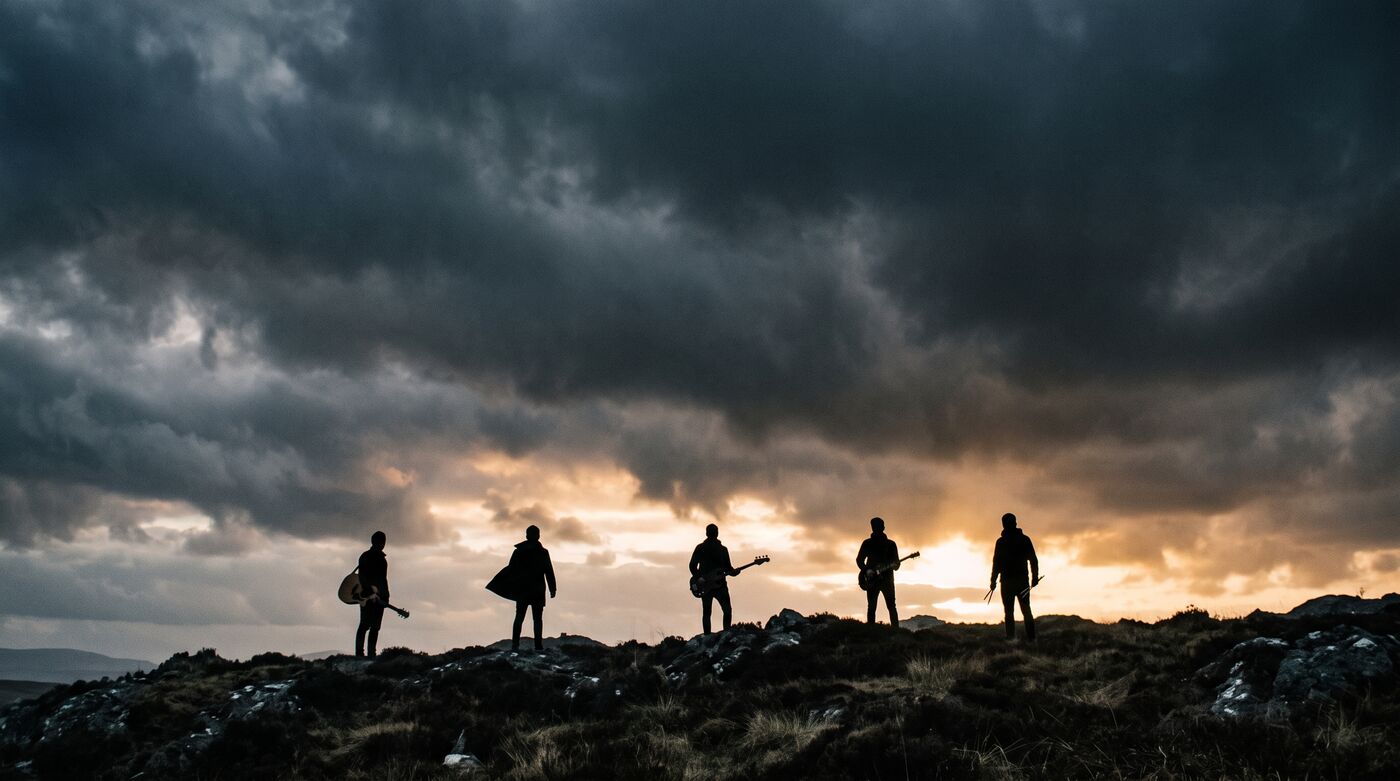Five silhouetted figures with instrument cases standing on a ridge under a storm sky, a gold break of dawn behind them.